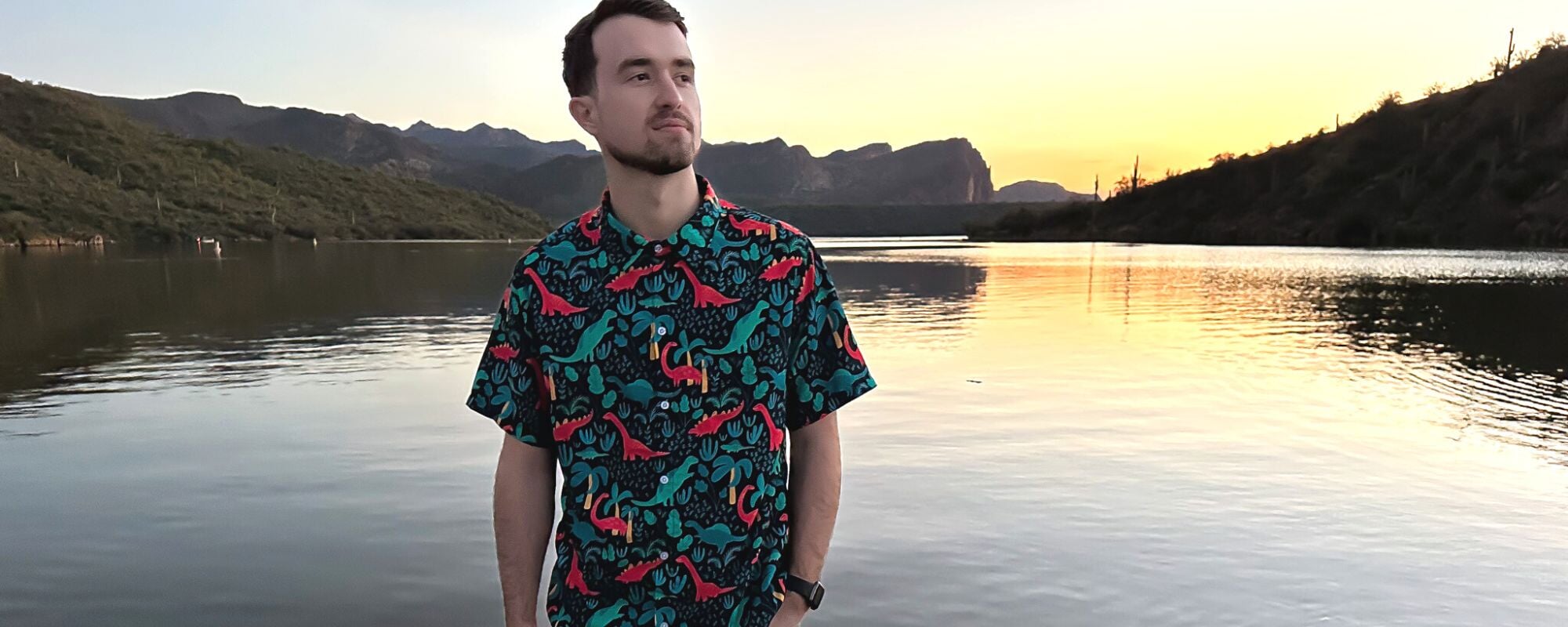 A man wearing a red and green dinosaur button down shirt stands on the shore of a lake during sunset. The tropical-themed shirt features colorful dinosaurs and foliage. 