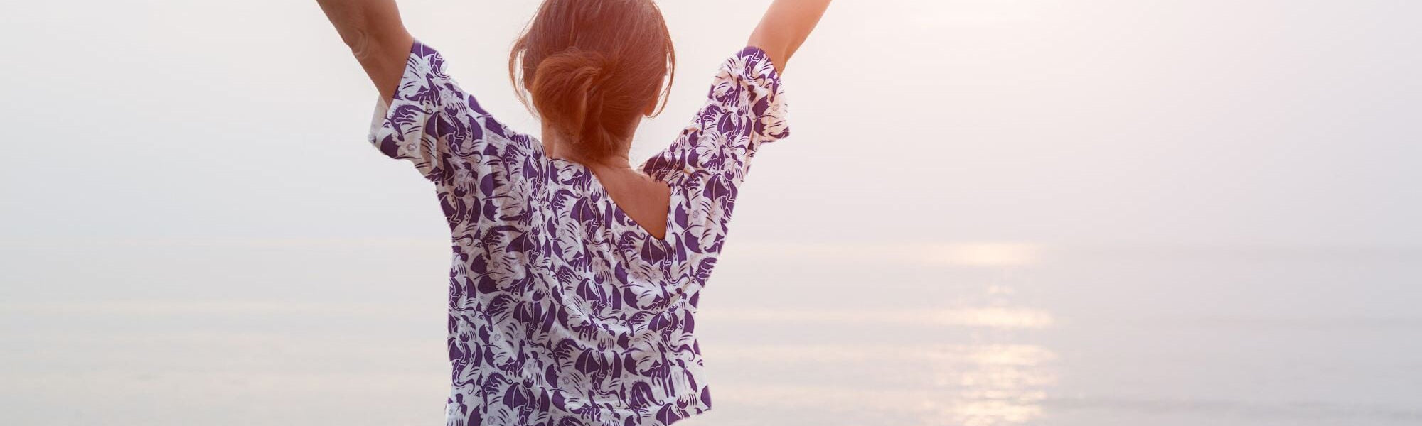 A girl wearing a fantasy printed shirt stands in the sun, looking out over the ocean. The unique design of the white button up shirt features fantastical purple dragons.