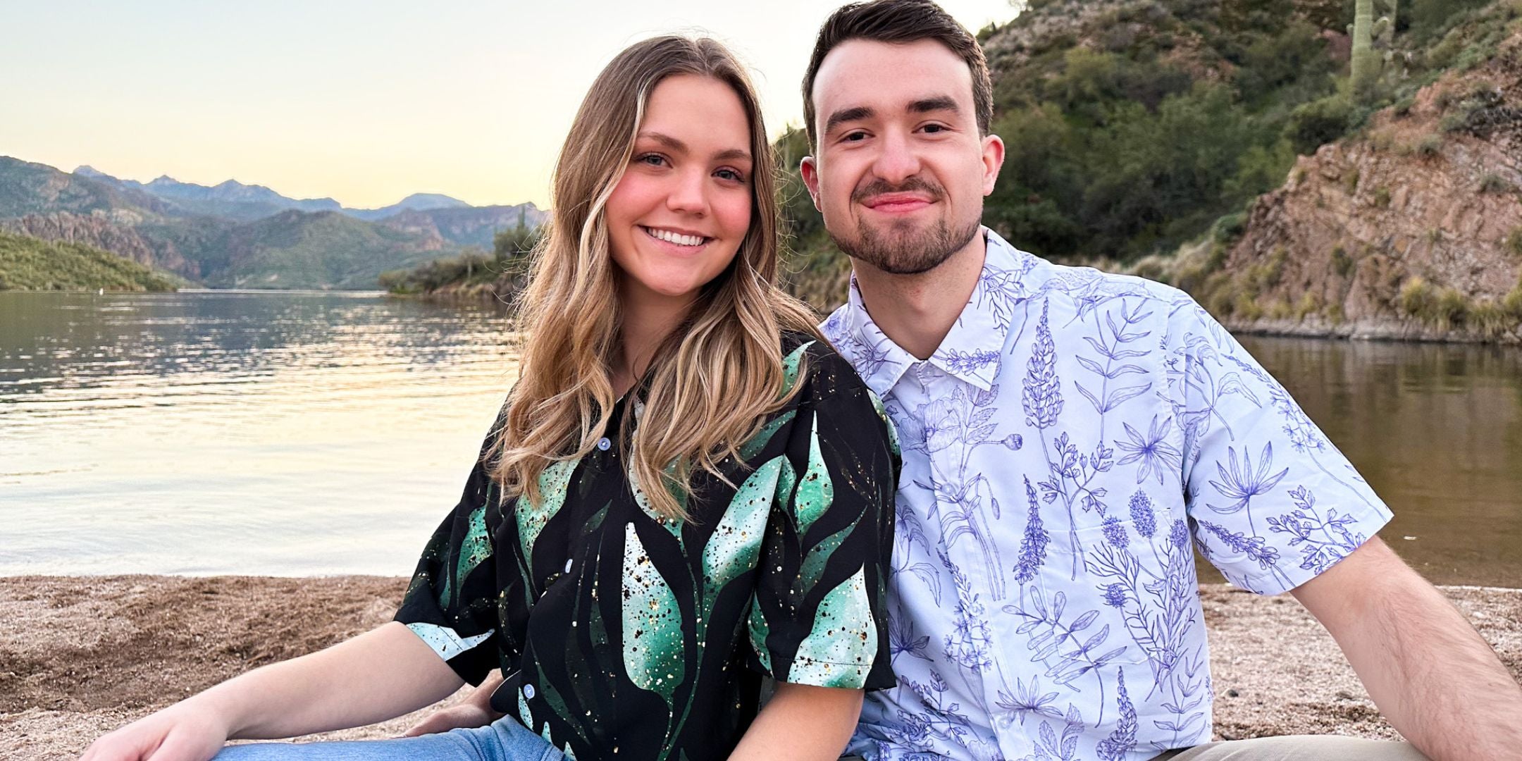 A couple sitting by a lake, both wearing stylish bontany button up shirts with botanical and plant prints. The man is wearing a white Hawaiian shirt with subtle leaves, while the woman is wearing a black botany shirt with green leaves that have mettallic glitter.
