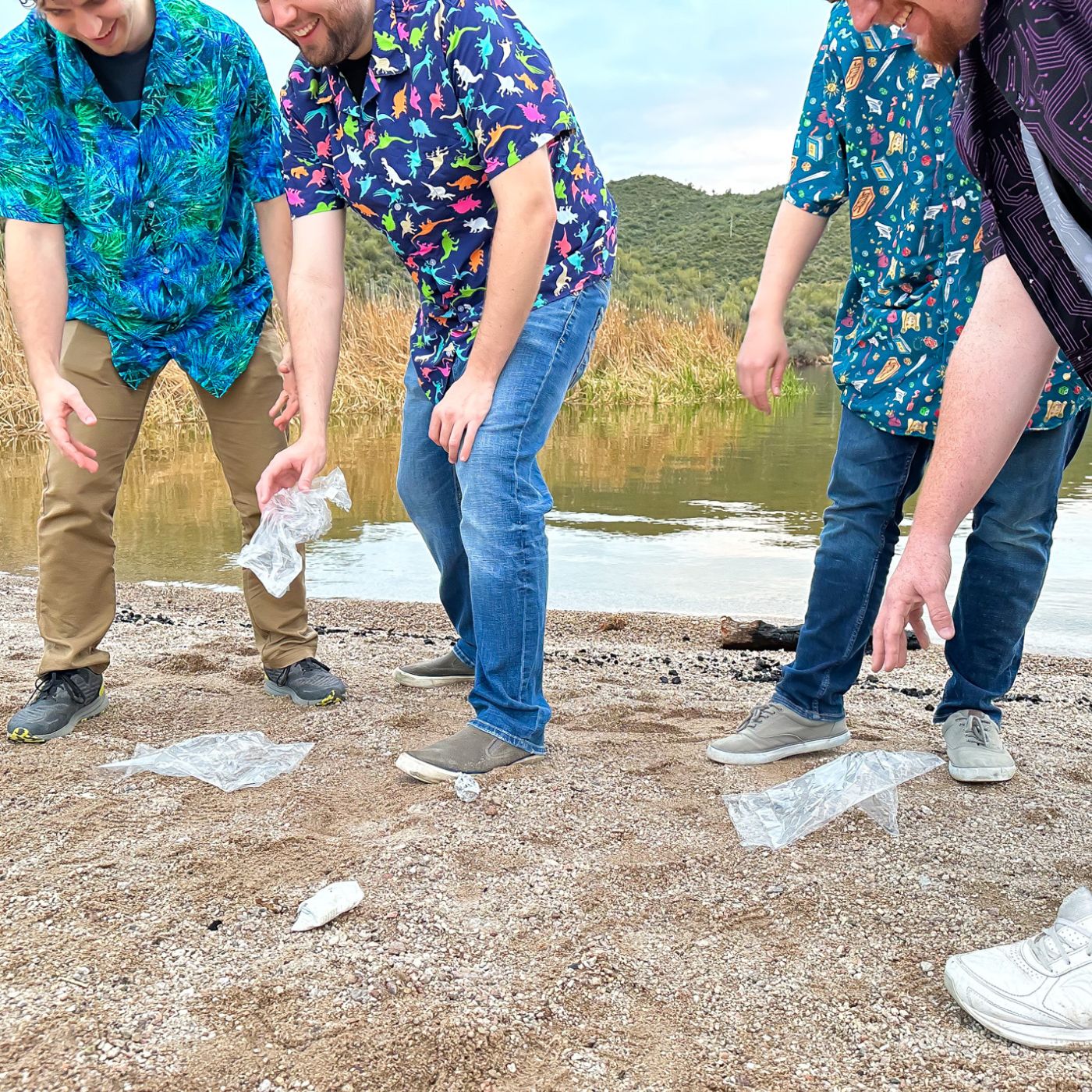 A group of people wearing colorful Geek Tropical button up shirts are picking up trash on a sunny tropical beach. The shirts feature fun and unique prints including dinosaurs, outer space, and superheroes. These eco-conscious individuals are doing their part to keep the beach clean while looking stylish and comfortable in their tropical-themed shirts.
