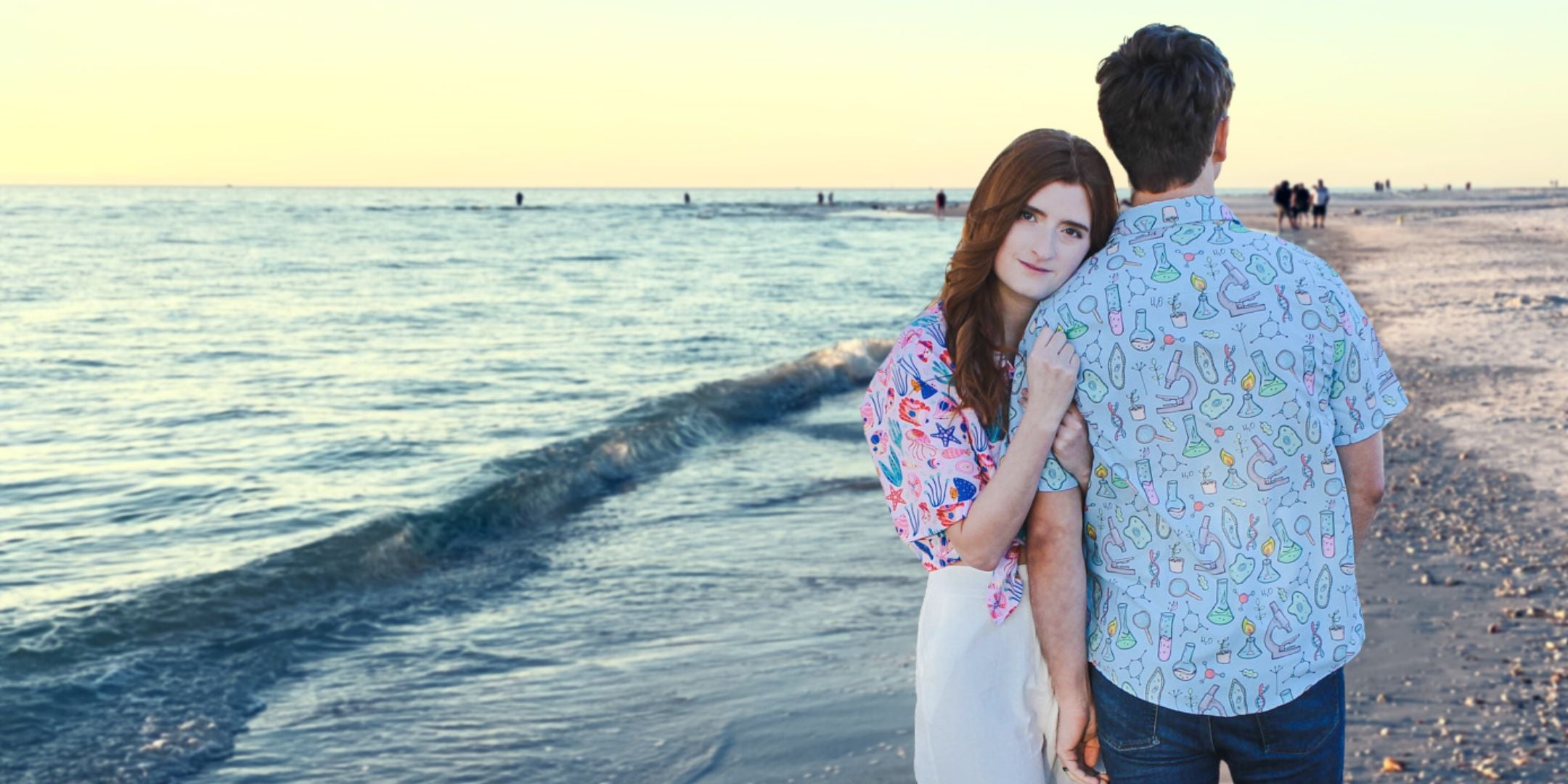 A couple wearing nerdy science button up shirts standing on the beach during a beautiful sunset. They are holding hands and smiling at each other while the waves are crashing behind them. The man's shirt has a pattern of DNA strands and the woman's shirt has a print of the periodic table.