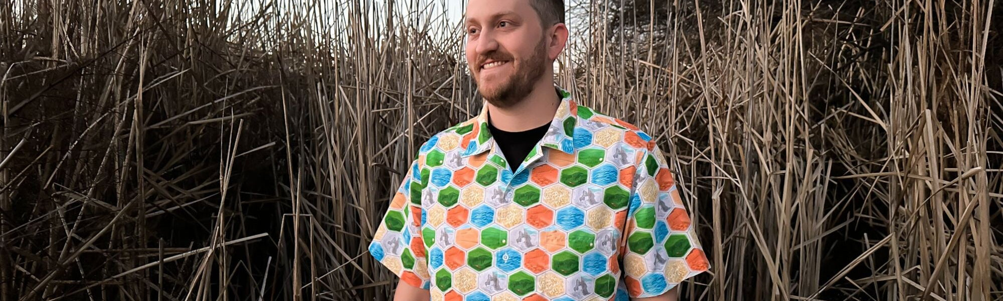 A man wearing a unique and colorful board game button up Hawaiian shirt with a hexagon tiles print. This nerdy shirt is perfect for any geeky outfit and will make you stand out in any crowd.