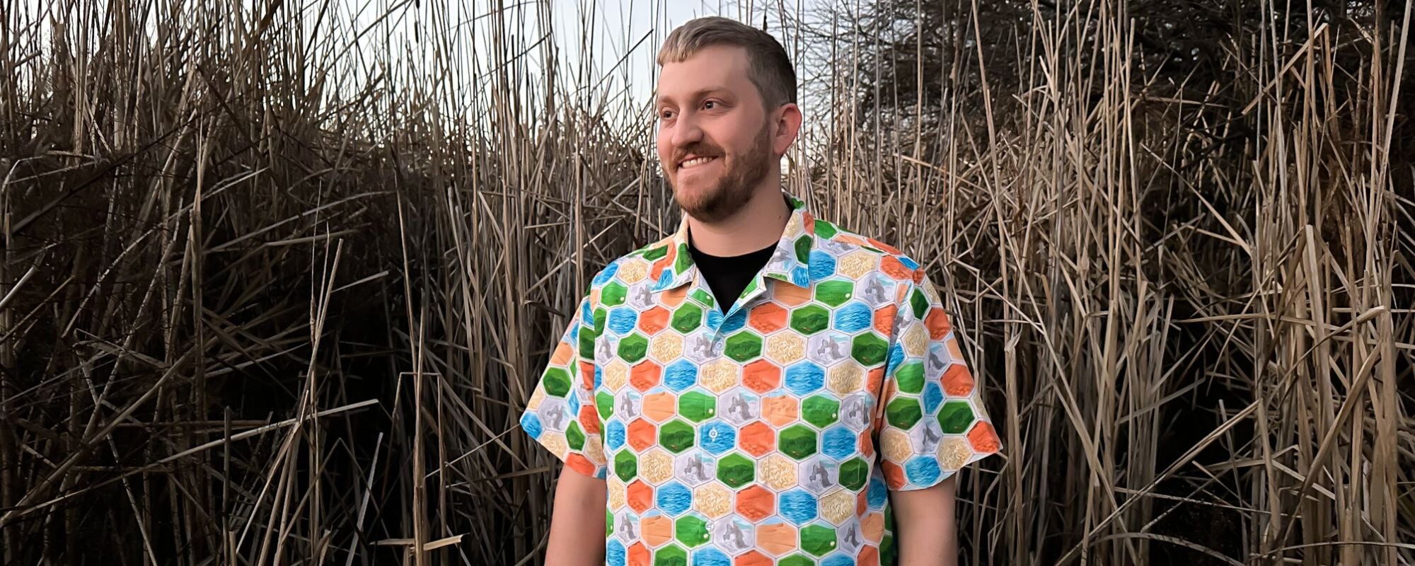 A man wearing a unique and colorful board game button up Hawaiian shirt with a hexagon tiles print. This nerdy shirt is perfect for any geeky outfit and will make you stand out in any crowd.