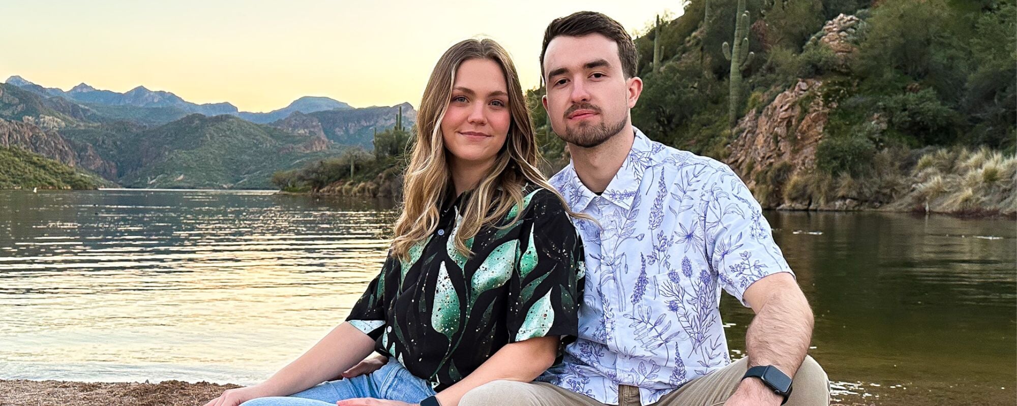 A couple sitting by a lake, both wearing stylish bontany button up shirts with botanical and plant prints. The man is wearing a white Hawaiian shirt with subtle leaves, while the woman is wearing a black botany shirt with green leaves.