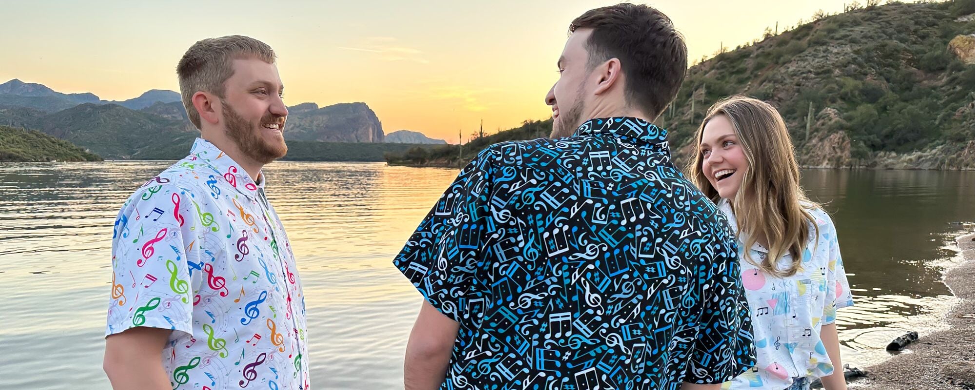 Three friends wearing their favorite geeky music themed button up shirts stand together at the edge of a lake at sunset. These shirts are perfect for music festivals or casual hangouts with fellow music enthusiasts.