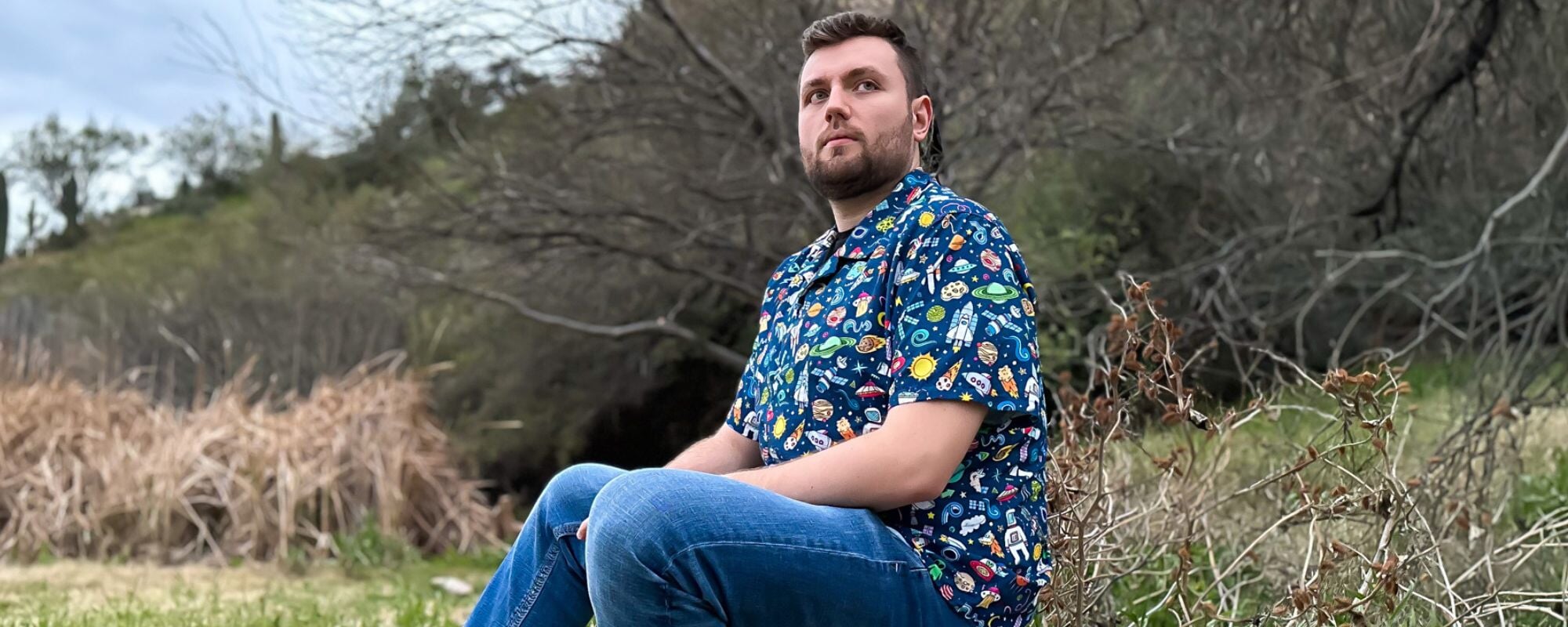 A man sits outdoors wearing his blue colorful outer space cartoon button down shirt. The shirt features an array of colorful cartoon planets, stars, and spaceships on a light blue background. 