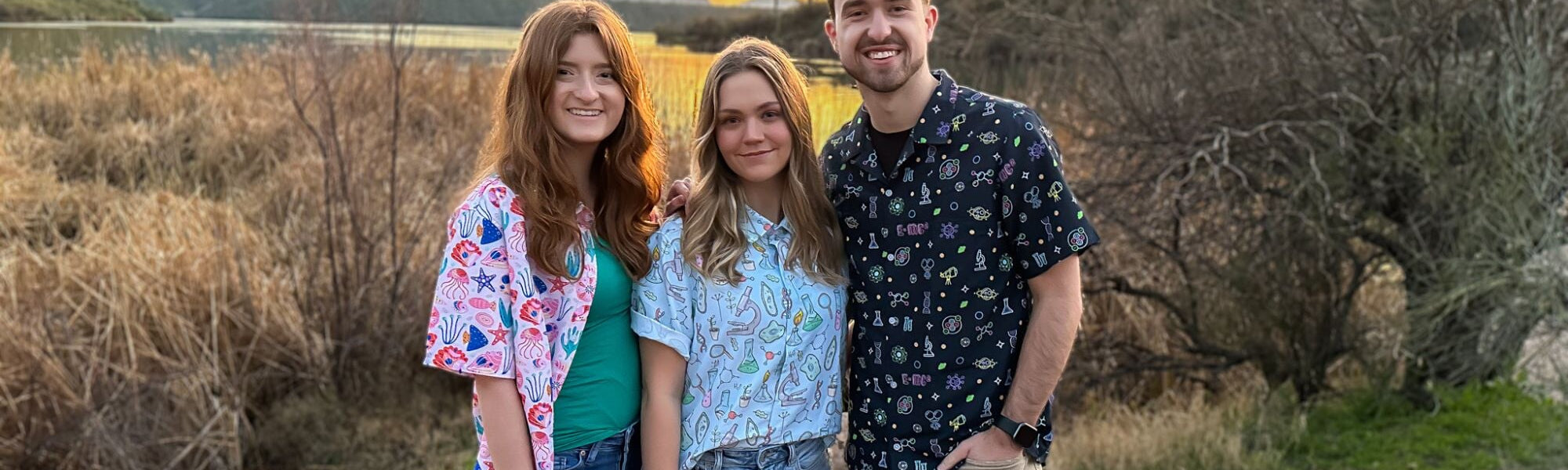 Three people wearing science themed button up shirts at a lake during sunset. The first person is wearing marine biology designs, the second person is wearing a bio-engineering button down shirt, and the third person is wearing a dark blue science shirt.