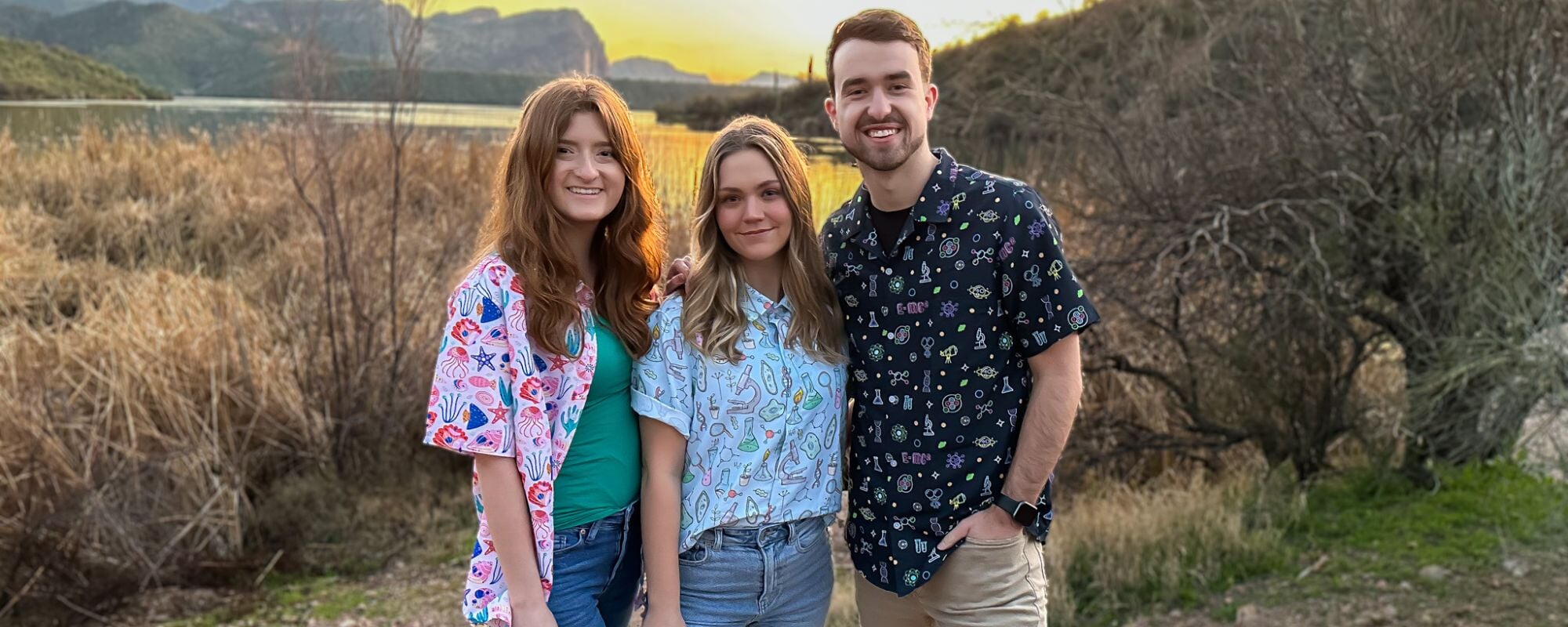 Three people wearing science themed button up shirts at a lake during sunset. The first person is wearing marine biology designs, the second person is wearing a bio-engineering button down shirt, and the third person is wearing a dark blue science shirt.