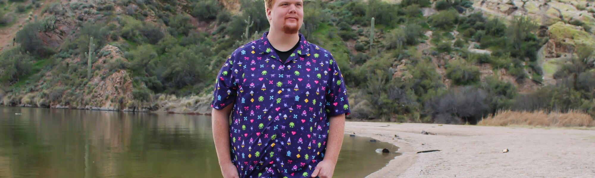 man standing on a sandy desert beach, wearing a vibrant purple shirt with classic video game arcade graphics printed all over it. The shirt features iconic video game characters and graphics in bright and bold colors