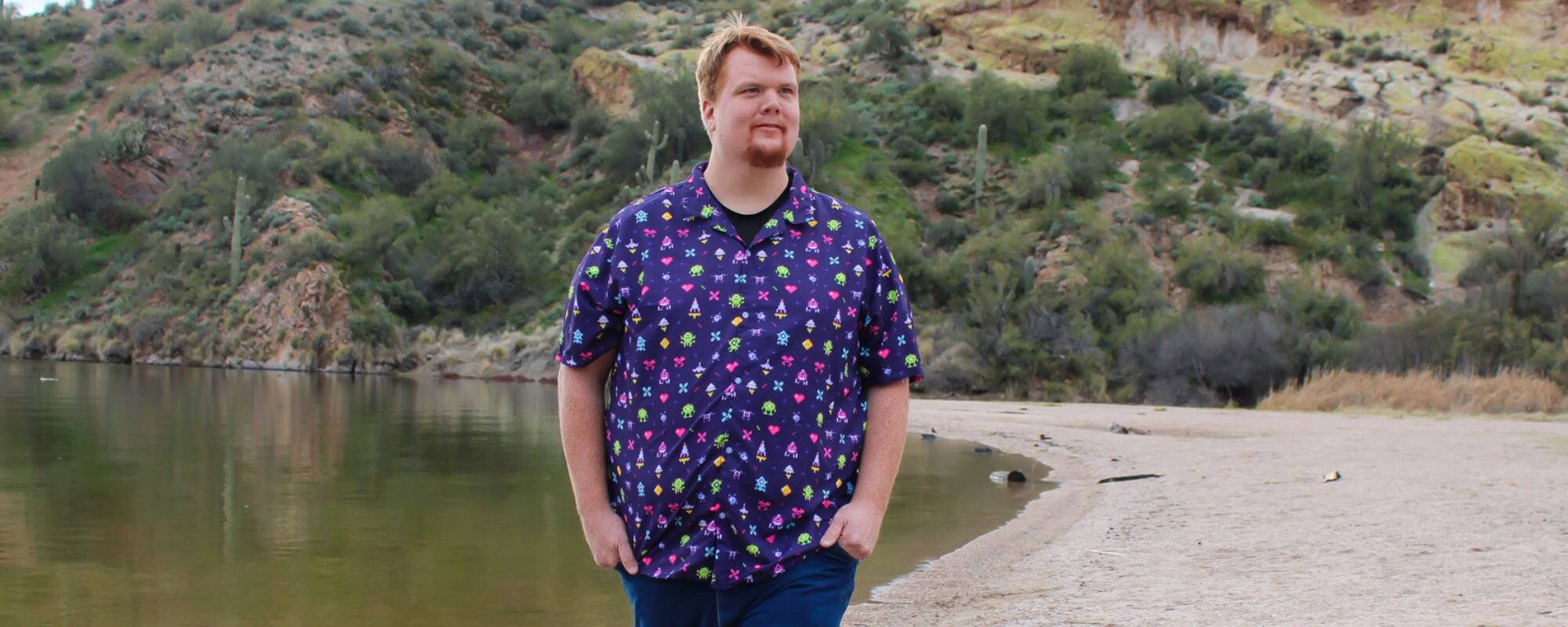 man standing on a sandy desert beach, wearing a vibrant purple shirt with classic video game arcade graphics printed all over it. The shirt features iconic video game characters and graphics in bright and bold colors