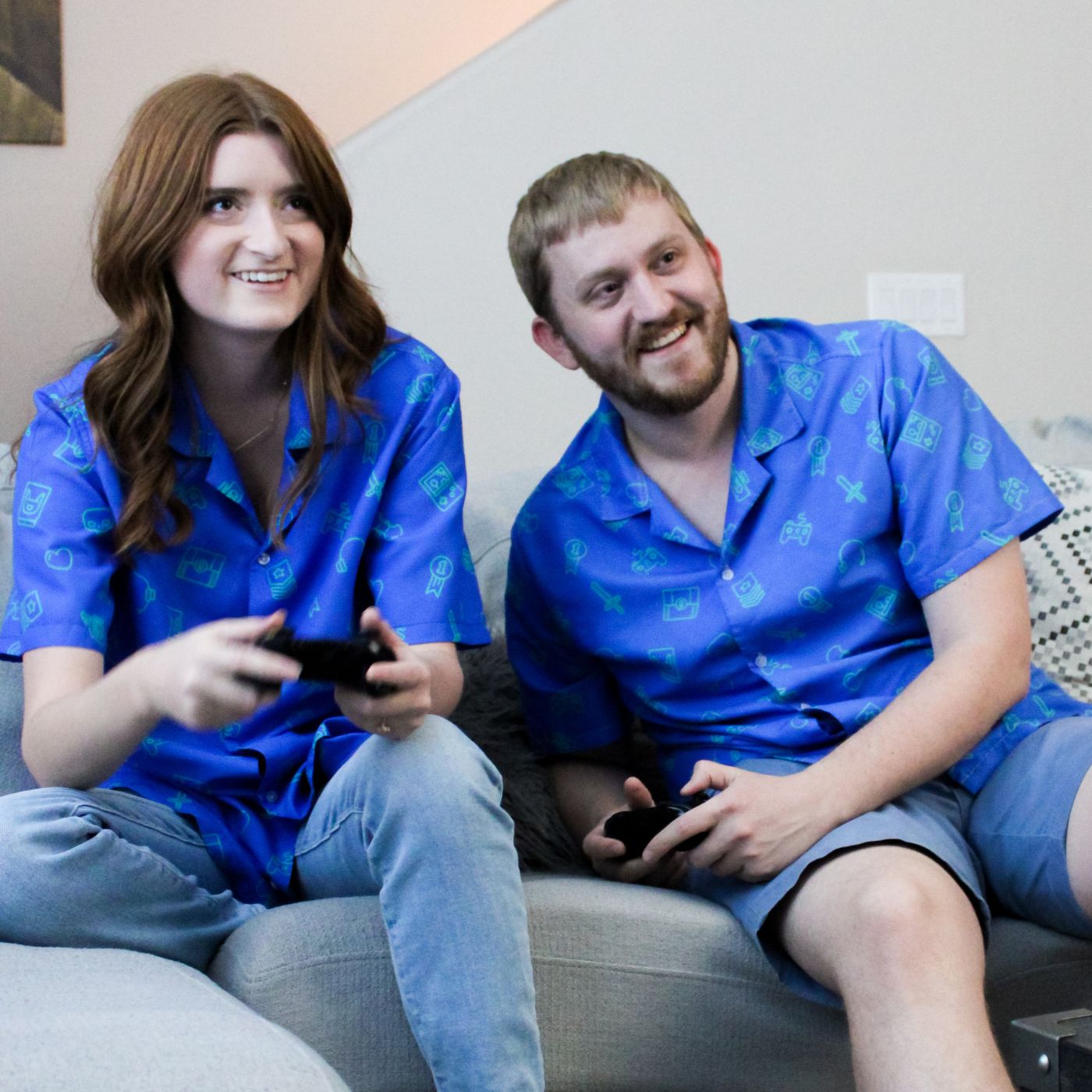A man and woman are sitting with gaming controllers wearing matching video game button up shirts. The prints have gamer icons typically seen in a video game.