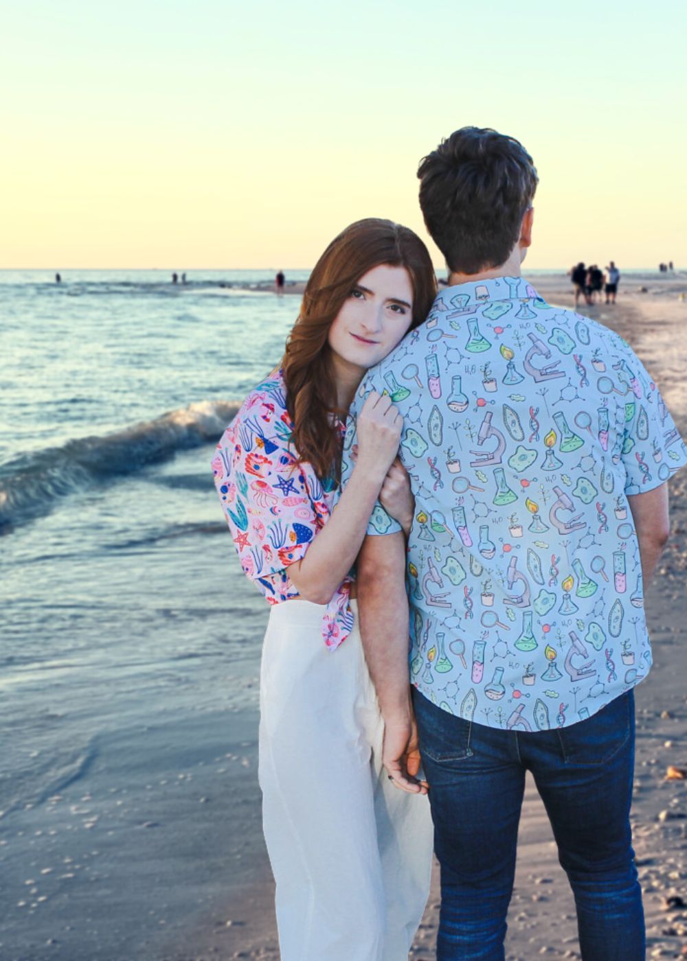 A couple wearing nerdy science button up shirts standing on the beach during a beautiful sunset. They are holding hands and smiling at each other while the waves are crashing behind them. The man's shirt has a pattern of DNA strands and the woman's shirt has a print of the periodic table.