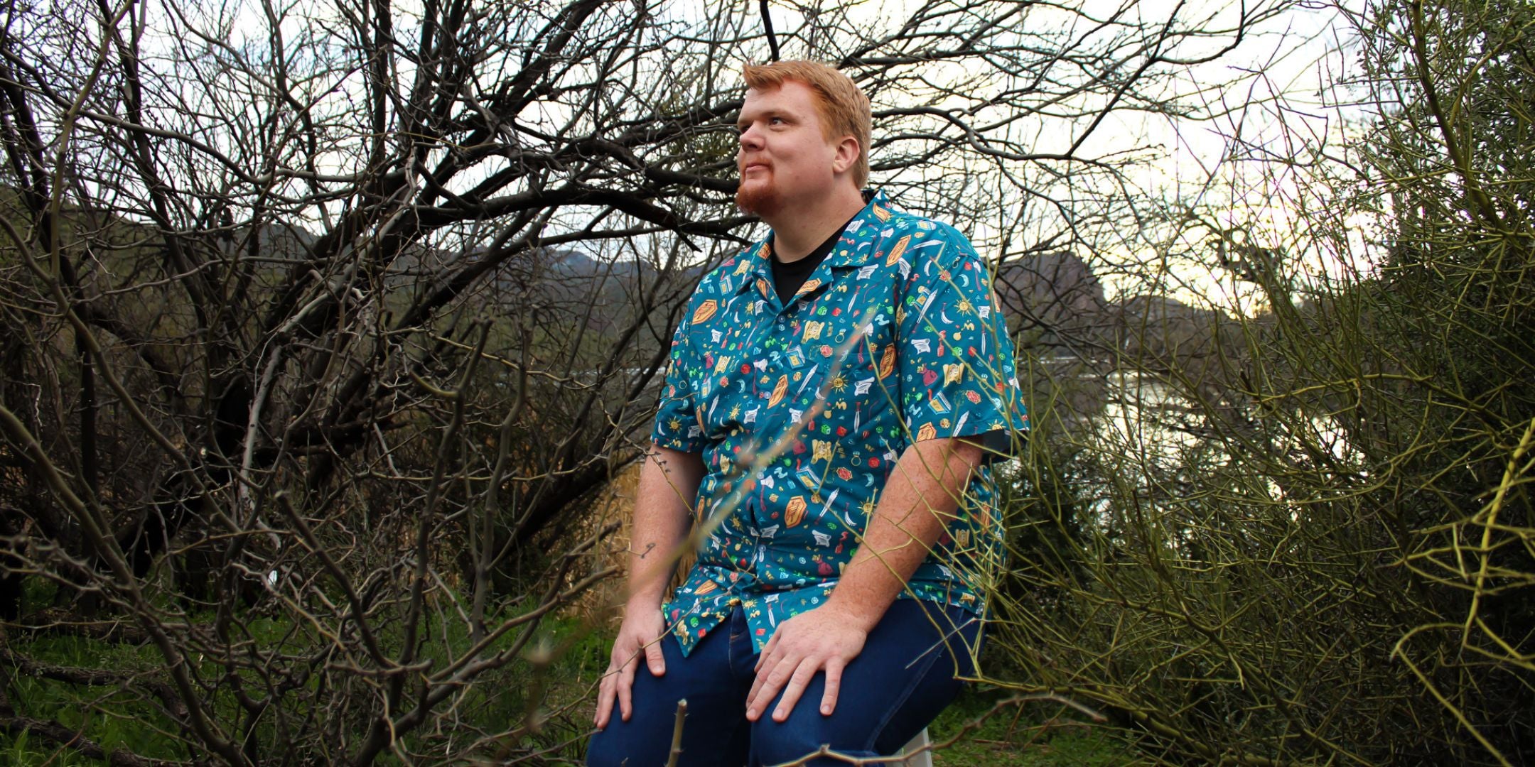 A red-headed man is standing in a lush forest wearing a RPG and dnd themed button-up adventure shirt. The shirt features dragons, swords, and other fantasy elements, perfect for any geeky outfit or D&D game night.