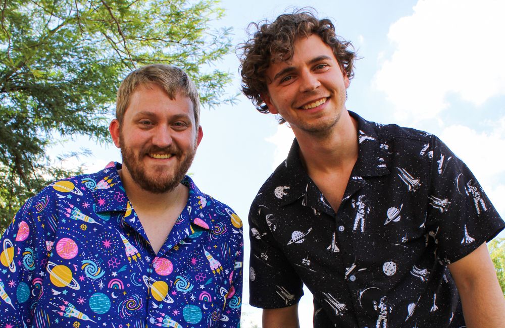 Two smiling men are wearing nerdy Hawaiian shirts with outer space prints. The cool button up shirts add a touch of geeky clothing to their tropical clothing.